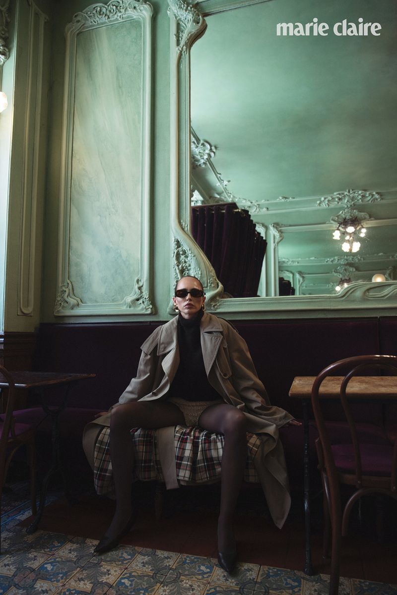 A stylish adult female sits on a bench in an indoor setting with a muted green wall, mirror, and tables in a fashion editorial.