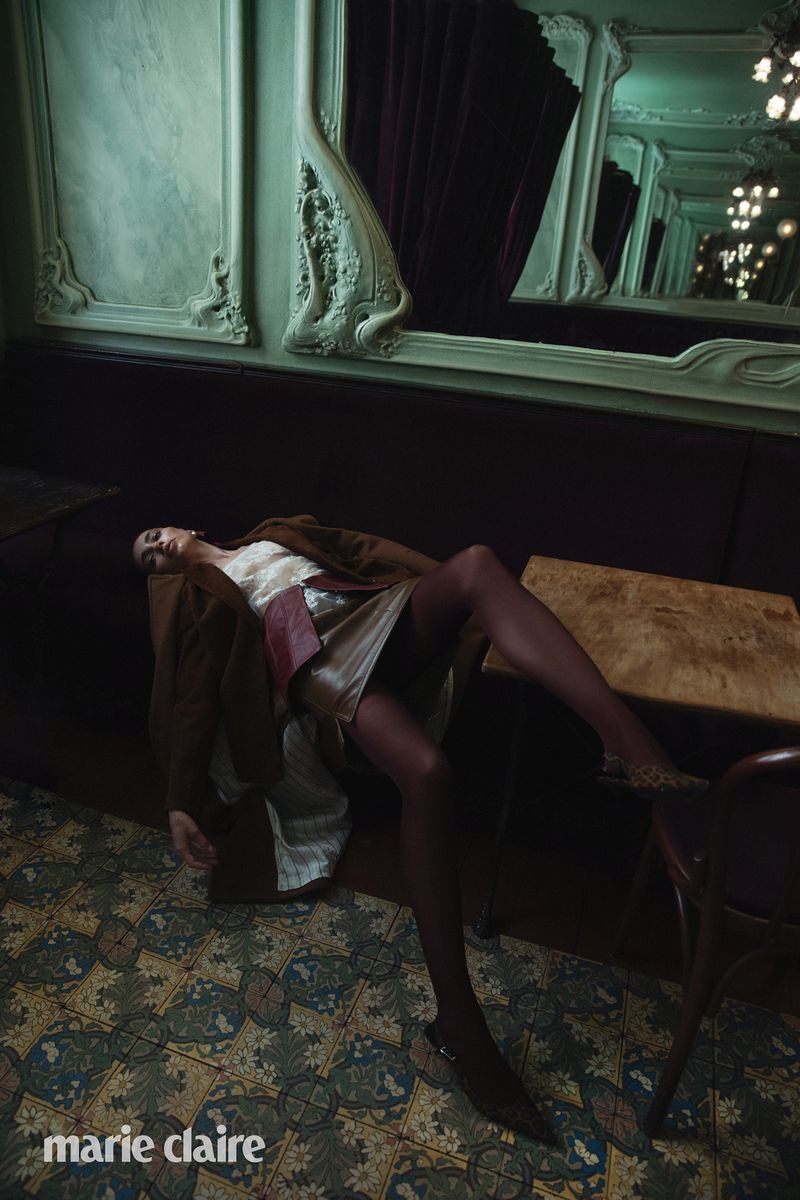 A woman lies diagonally across a settee in a brown coat and nude hose, in an indoor setting with tiled floors and a dark wooden table.