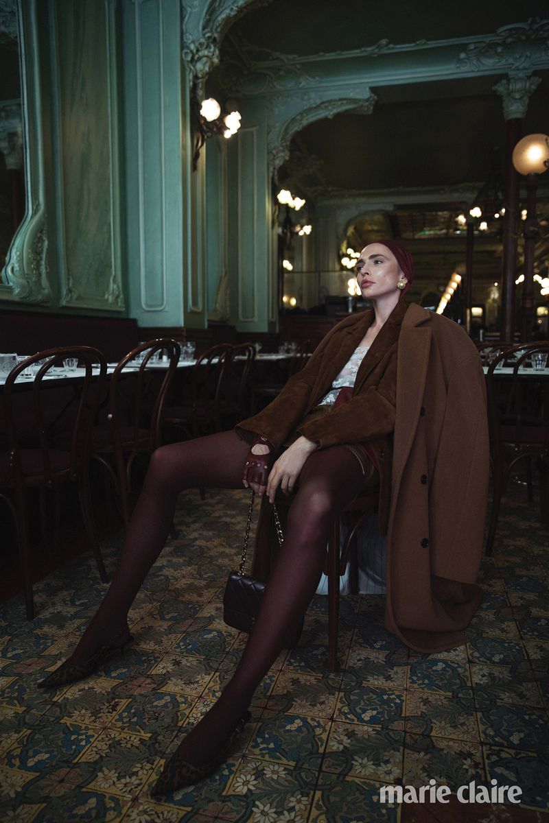 A woman in a brown coat and burgundy tights sits in a vintage cafe, holding a small purse. Classic bistro chairs and lighting.