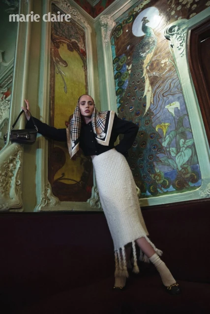 Fashion editorial shot of a posed woman in a black jacket, white skirt, scarf, and handbag, standing against an ornate peacock-themed wall.