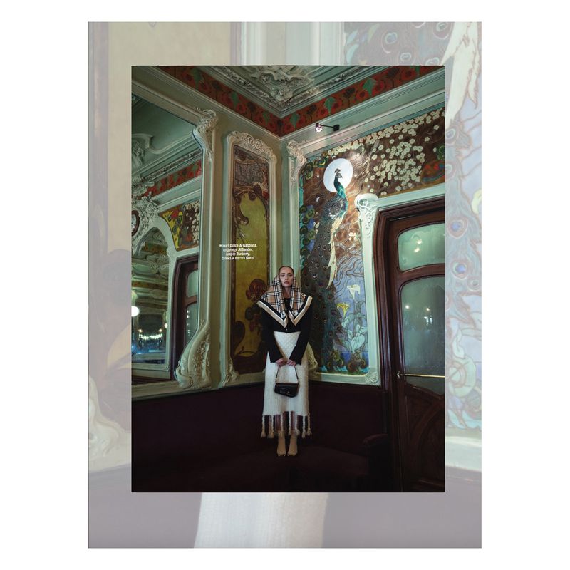 Indoor shot of a woman in a black blazer and long cream skirt, holding a black purse, in front of decorative wall panels.