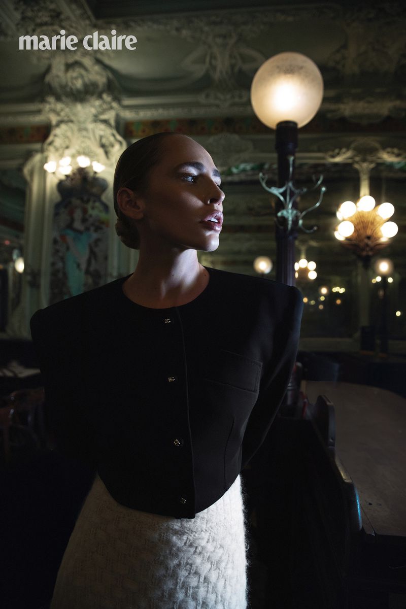 A woman in a black jacket and white skirt poses indoors with decorative lamps and ornate architecture for a fashion editorial.