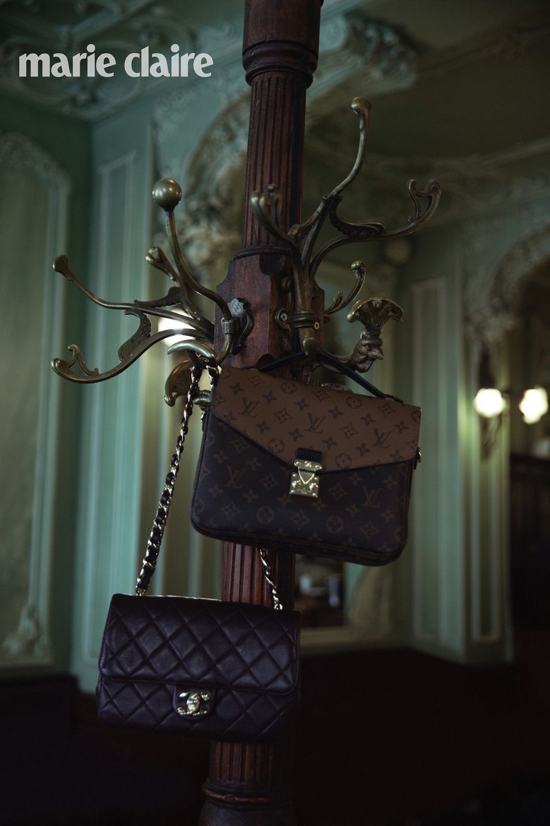 Two luxury handbags hang on a vintage coat rack inside an ornate building. The bags are brown and black with gold hardware.