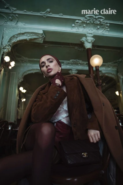 Fashion editorial image in a European cafe featuring a woman wearing a brown coat, headscarf, and Chanel purse in an indoor setting.
