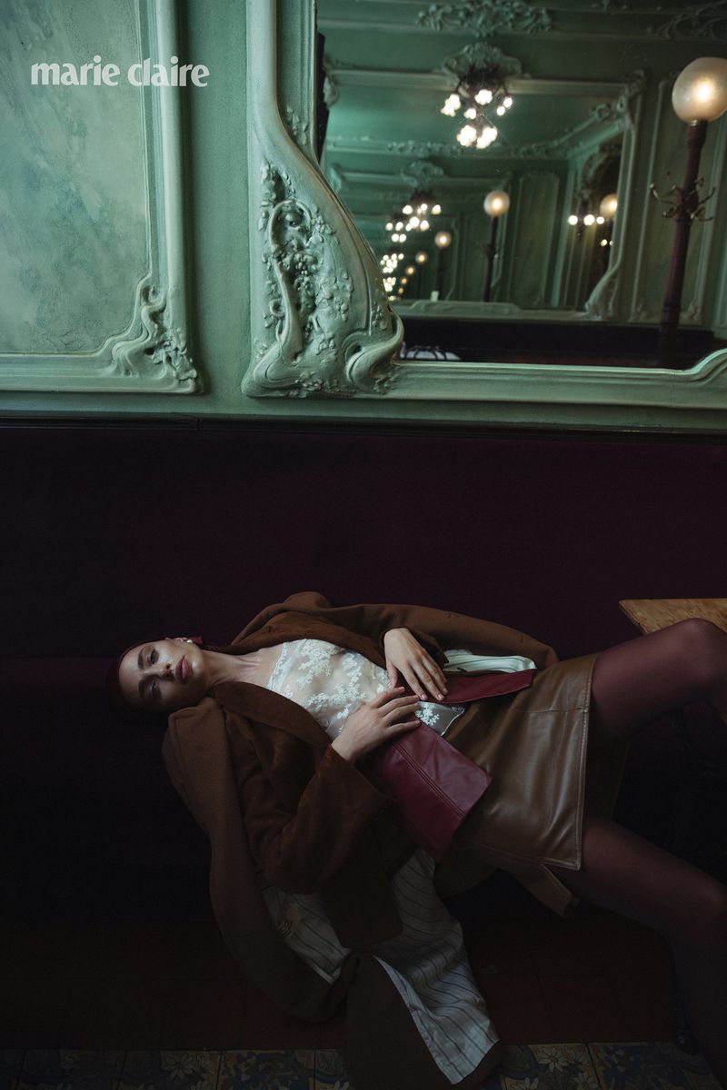Woman in brown coat lies on a maroon couch with ornate architectural details and mirrored wall in the background with globe lights.