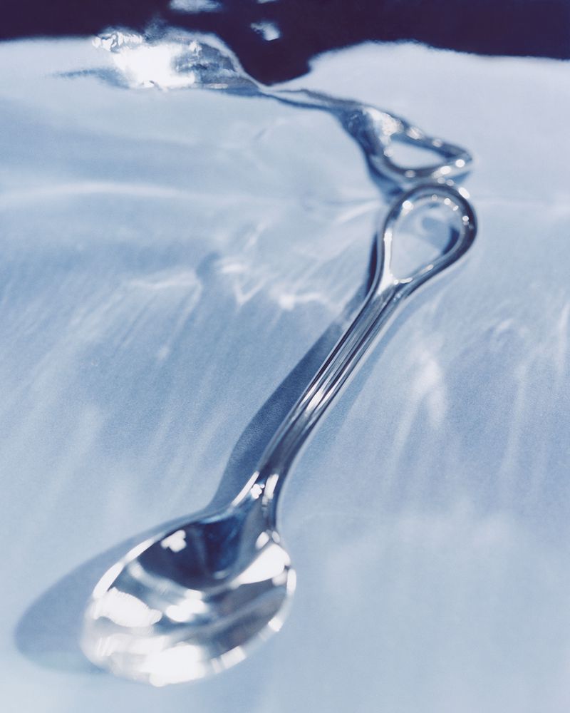 Shiny silver spoon rests on a blue surface with refracting light patterns, suggesting a still life with a minimalist aesthetic.