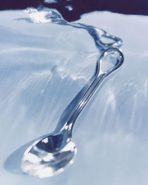 Shiny silver spoon rests on a blue surface with refracting light patterns, suggesting a still life with a minimalist aesthetic.