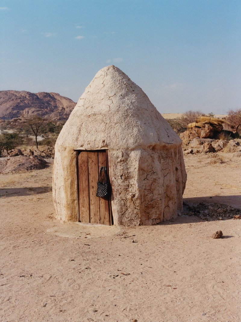 Mud hut with wooden door in arid African landscape under blue sky. A woven bag hangs near the entrance of the earthen structure.