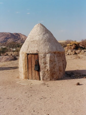 Mud hut with wooden door in arid African landscape under blue sky. A woven bag hangs near the entrance of the earthen structure.