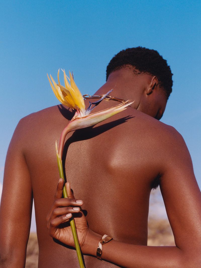 Back view of a shirtless dark-skinned adult male with an afro, holding a yellow bird of paradise flower against a clear blue sky.