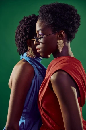 Two Black women in profile against a green backdrop, one wearing a draped red dress and heart tassel earrings, the other in blue.