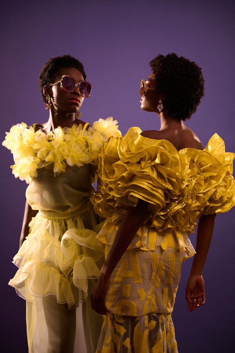 Two Black models posing in voluminous yellow ruffled couture dresses and statement sunglasses against a solid purple backdrop.