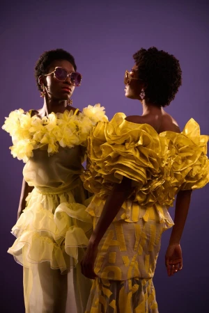 Two Black models posing in voluminous yellow ruffled couture dresses and statement sunglasses against a solid purple backdrop.