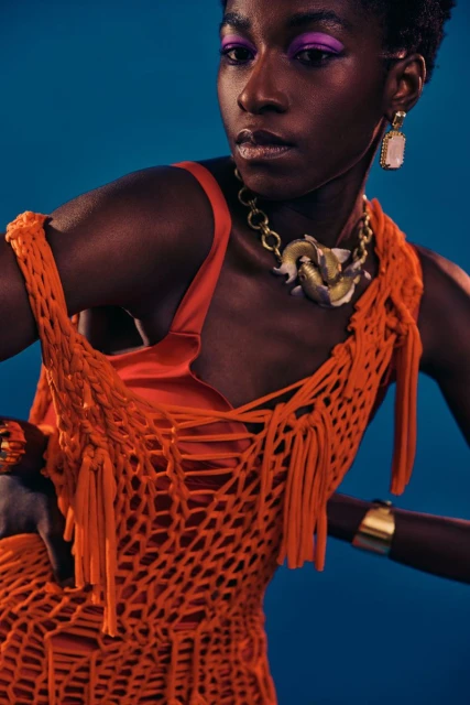 A dark-skinned woman with short hair poses wearing an orange fringe dress, gold jewelry, and vibrant purple eyeshadow against blue.