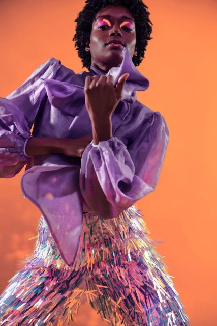 Fashionable person with dark skin and curly hair poses wearing a ruffled lavender blouse and iridescent tinsel pants against an orange backdrop.