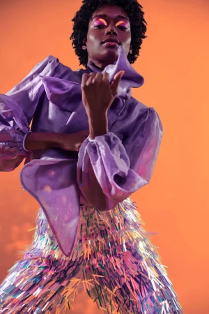 Fashionable person with dark skin and curly hair poses wearing a ruffled lavender blouse and iridescent tinsel pants against an orange backdrop.