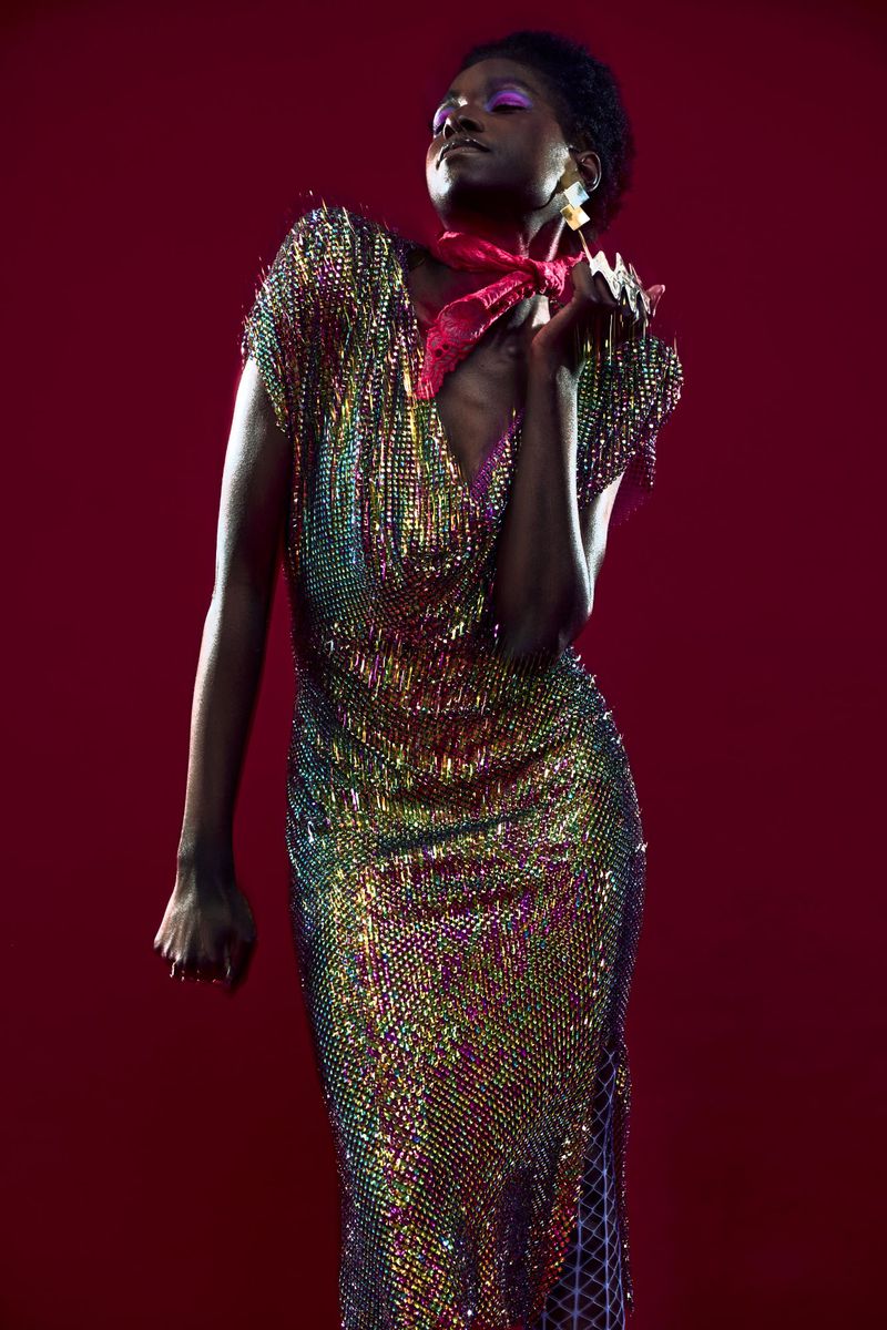 Elegant Black woman poses in a colorful shimmering gown with pink scarf, highlighting fashion and beauty against a deep red background.