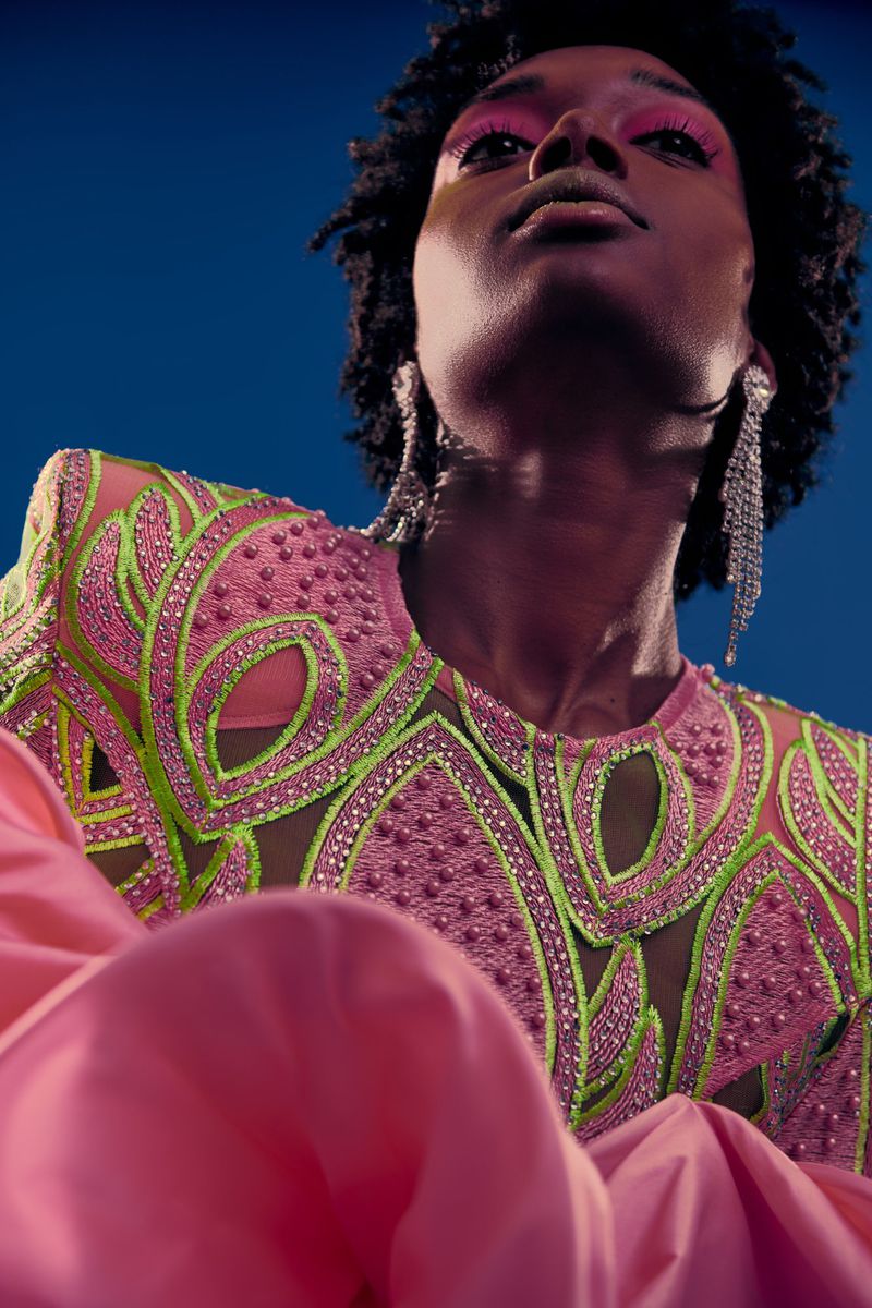 Portrait of an African American female model wearing a pink dress with green embroidery looking up against a dark blue background.