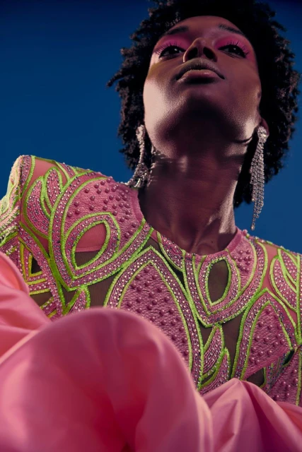 Portrait of an African American female model wearing a pink dress with green embroidery looking up against a dark blue background.