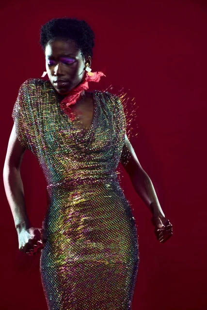 Black female fashion model in iridescent beaded dress poses against a red studio backdrop with magenta makeup and a red neck scarf.