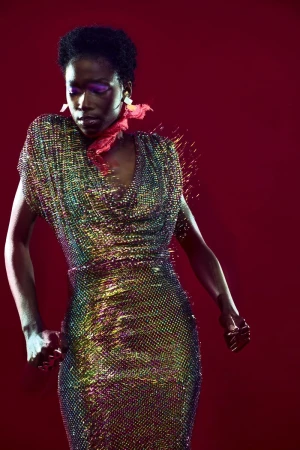Black female fashion model in iridescent beaded dress poses against a red studio backdrop with magenta makeup and a red neck scarf.