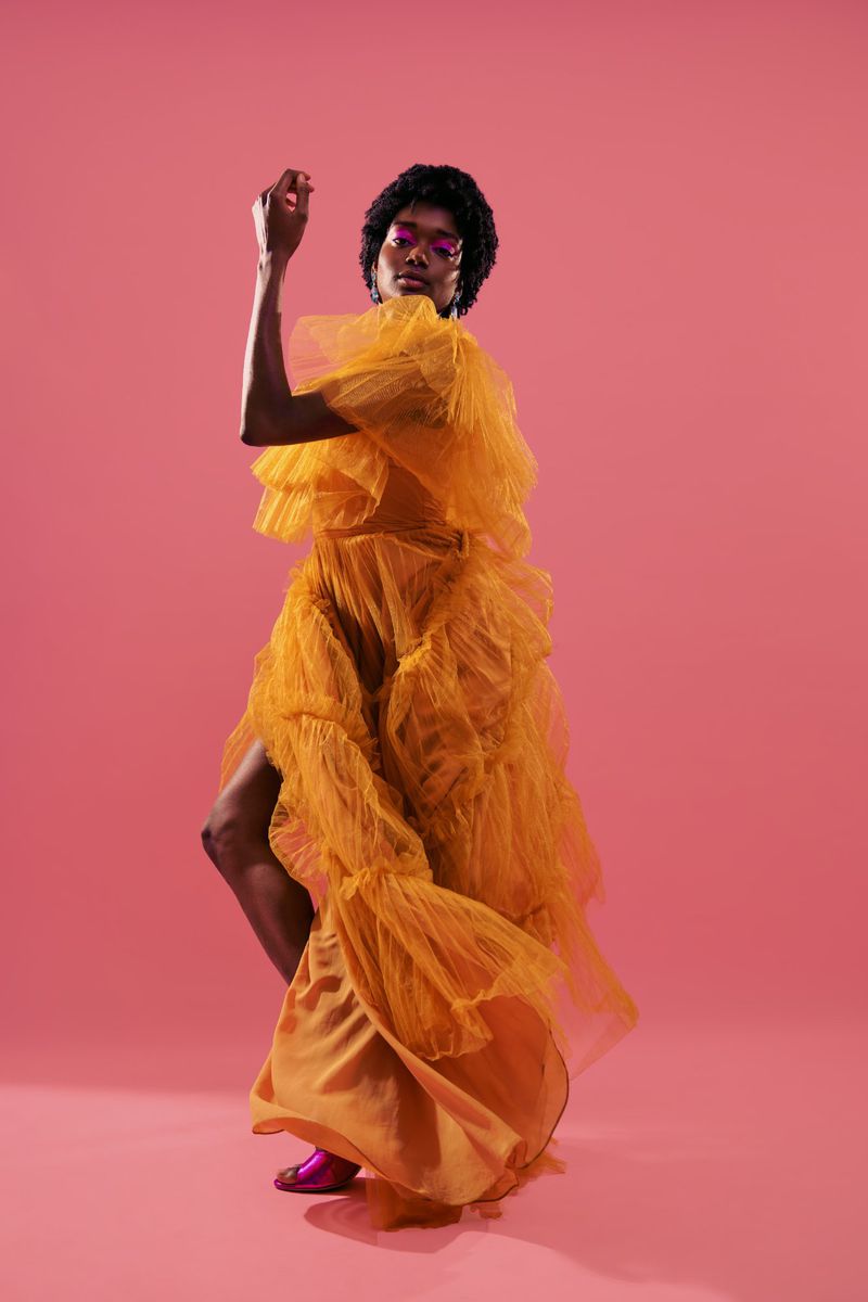 Woman in a golden ruffled gown poses against a pink background in a studio, showcasing a theatrical and fashionable look.