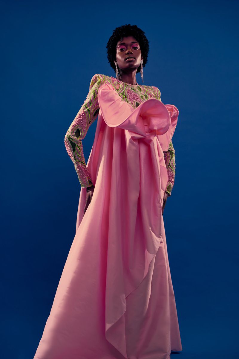 Woman in elaborate pink gown with floral embellishments and beaded sleeves poses against a solid blue background in a studio setting.