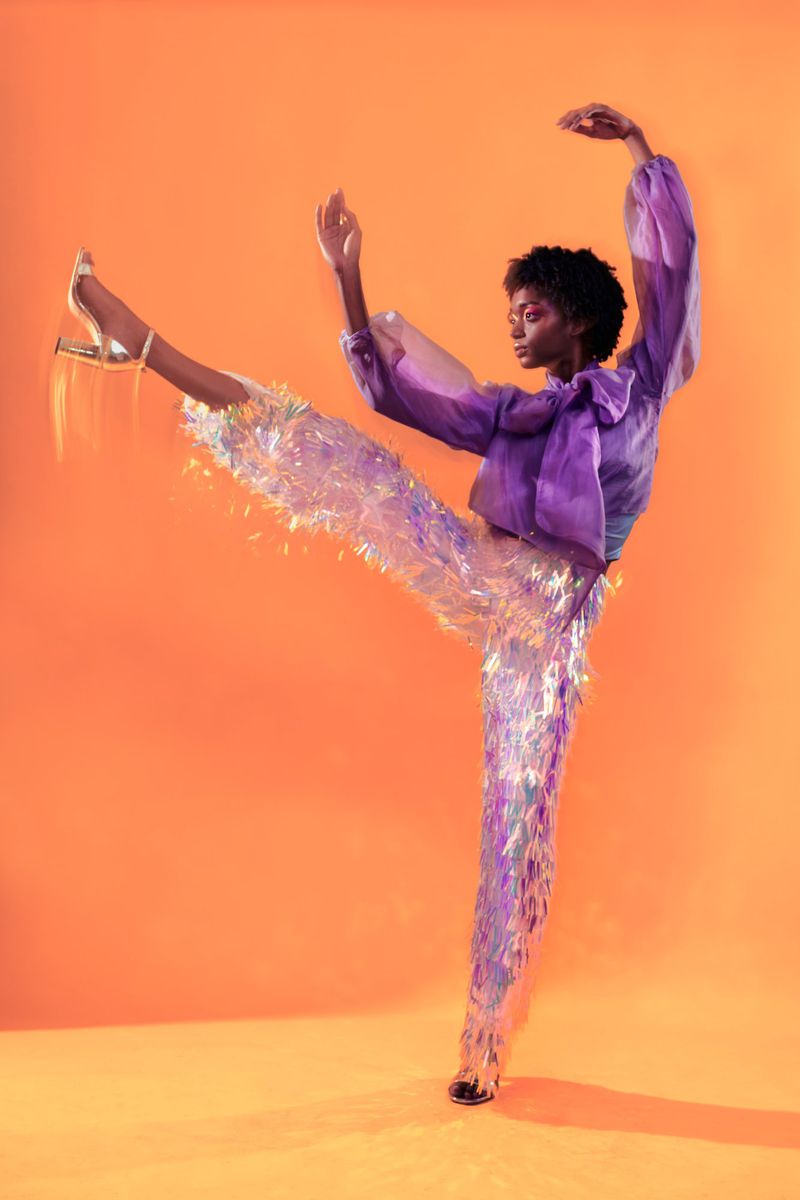 A striking full-body shot of an African American woman in motion, wearing a purple top and iridescent fringe pants against an orange backdrop.