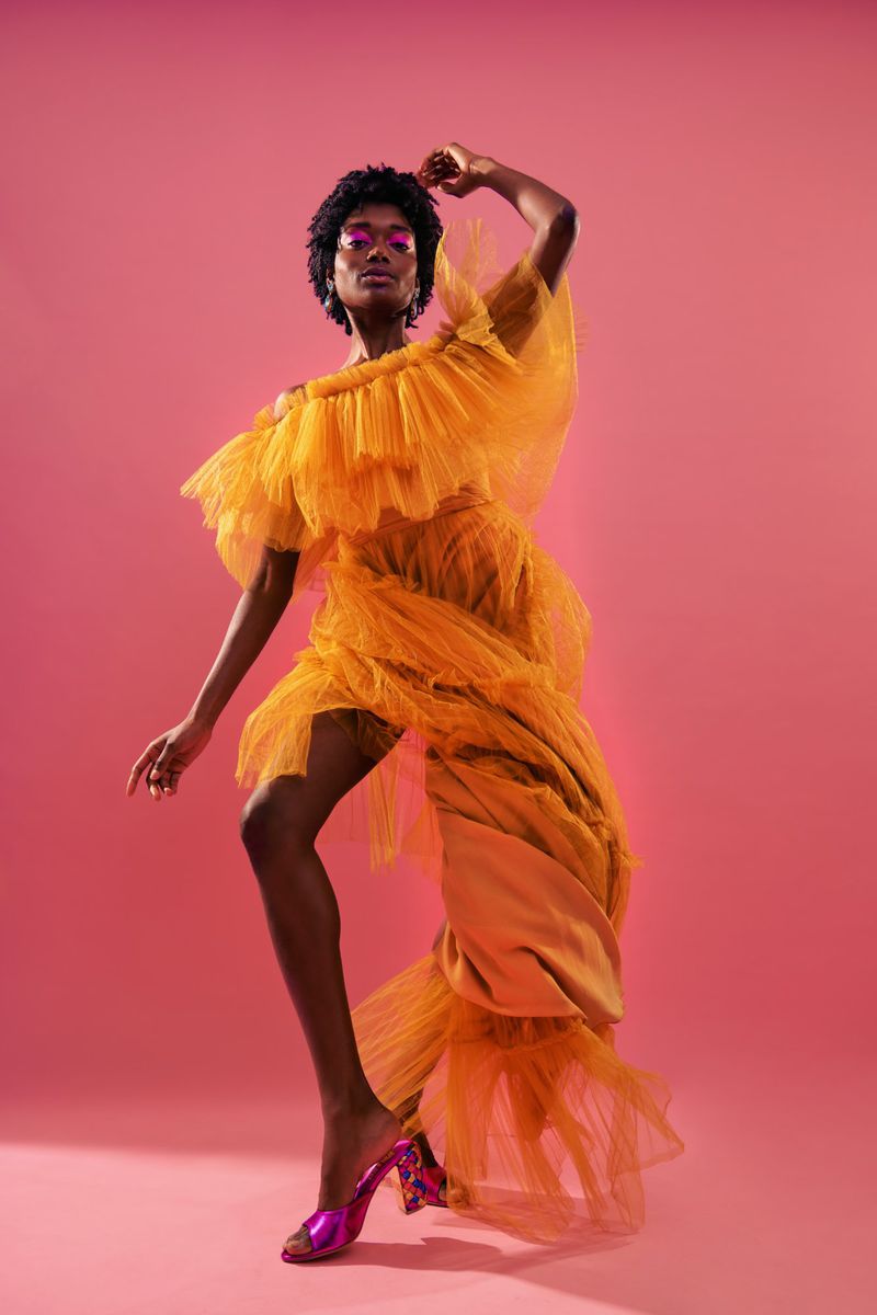 A woman with dark skin and short hair poses in a bright gold gown, with pink eyeshadow and matching heels, against a pink backdrop.