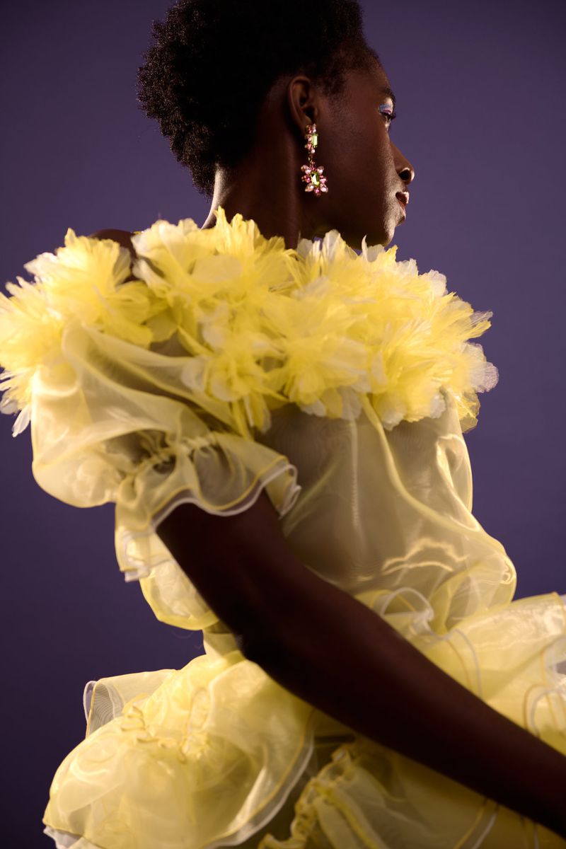 Elegant woman with dark skin in a light yellow dress with floral earrings poses against a purple background in a fashion setting.