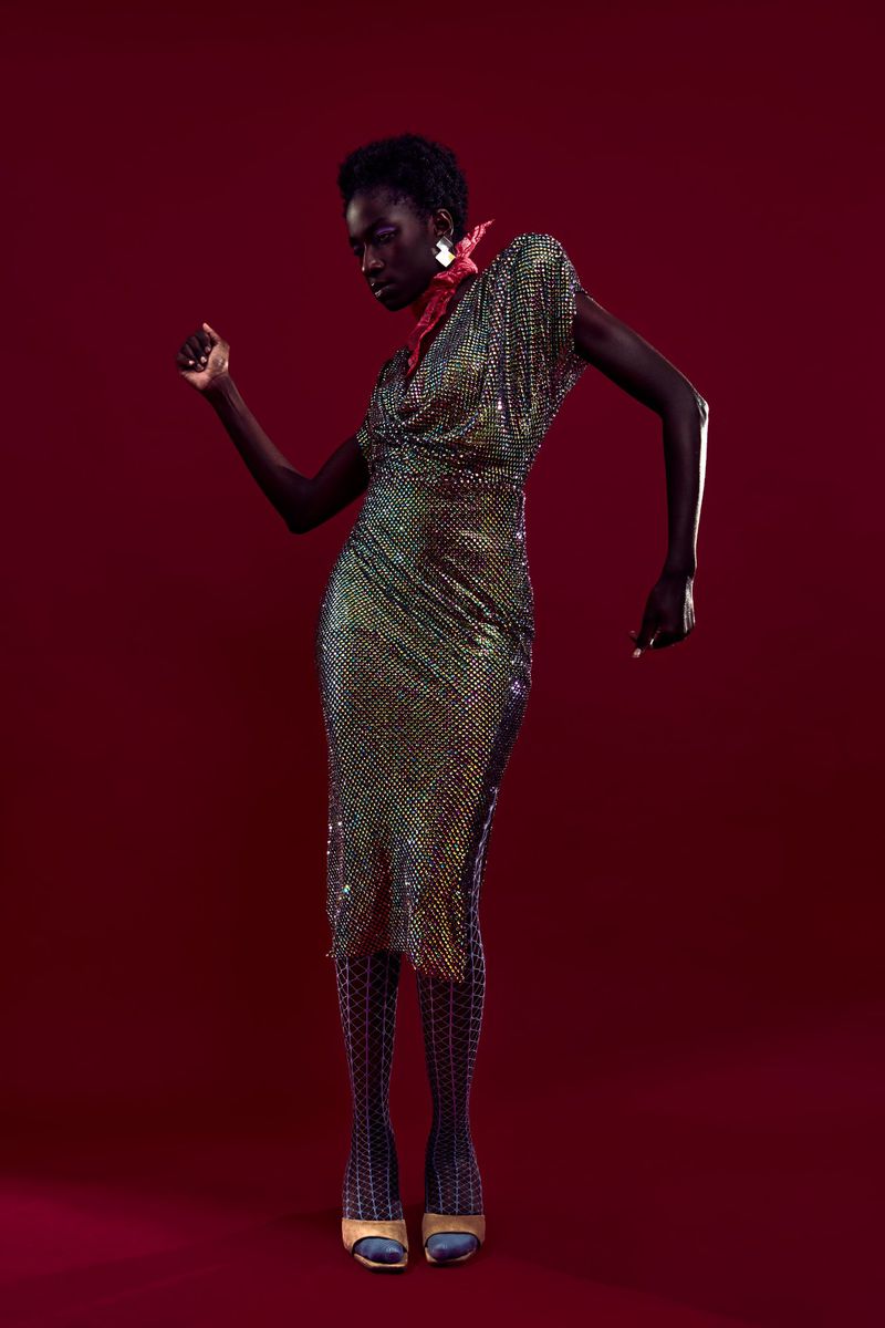 Elegant Black woman in iridescent chainmail dress poses in studio against a maroon background, fashion photography, chic style.