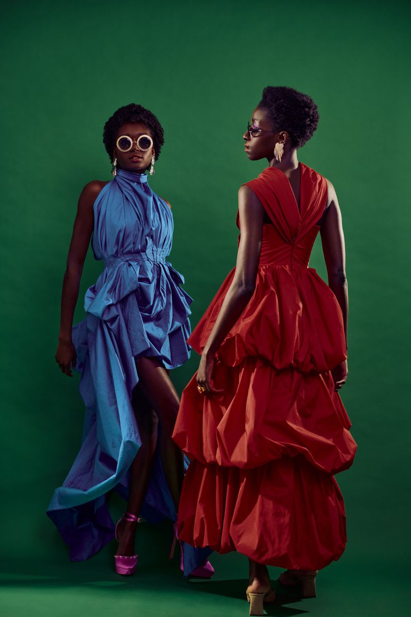 Two Black women in draped dresses pose against a green backdrop. One wears blue with pink heels, the other red with neutral heels.