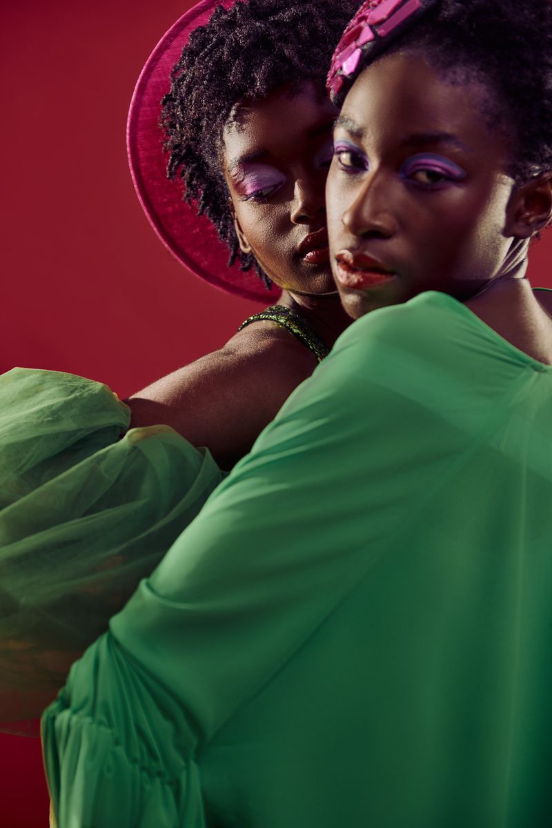 Two African American women pose in a studio portrait, one in a green top with a pink hat, against a red backdrop, showcasing fashion and beauty.