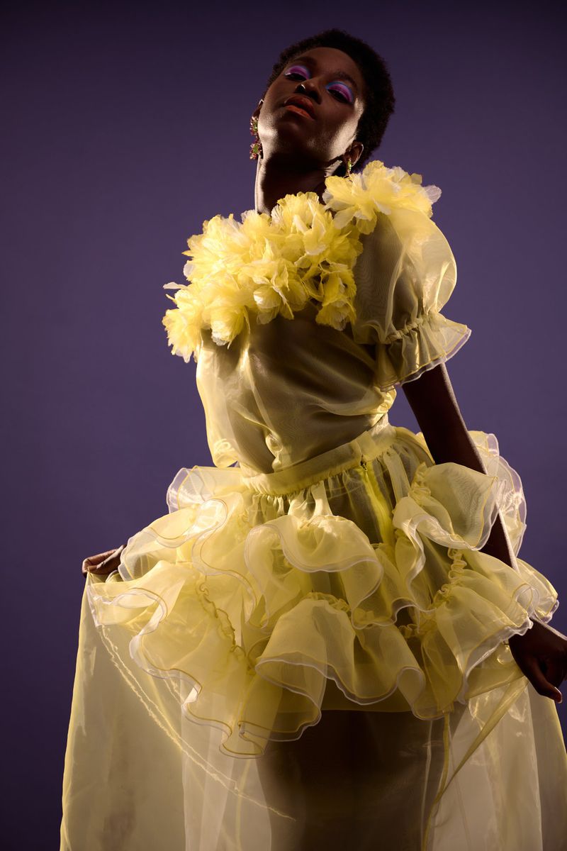 An African-American female model in a yellow tulle dress with purple eye makeup strikes a pose against a purple backdrop in studio lighting.