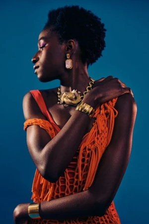 Stylish portrait of a Black woman wearing orange fringe top, gold jewelry on a blue backdrop in a fashion or beauty context.