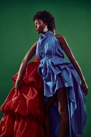 A Black woman posing in a draped blue halter dress and large statement earrings against a solid green studio backdrop.