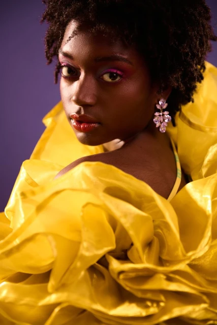 Close-up portrait of an African American woman with dark hair and pink makeup wearing large yellow ruffled clothing against a purple background.