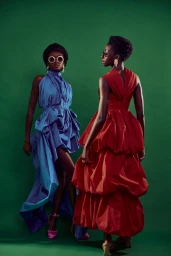 Two Black women in dramatic blue and red tiered gowns posing against a solid green seamless studio background.