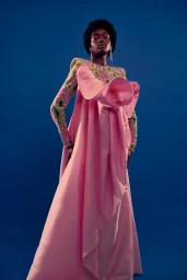 Black model posing in a voluminous floor-length pink gown with embroidered sleeves against a solid blue studio backdrop.