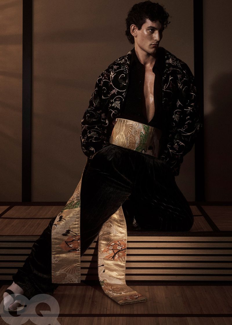 A model poses indoors wearing a black shirt and velvet pants with gold embroidered details, against a neutral background with soft lighting.