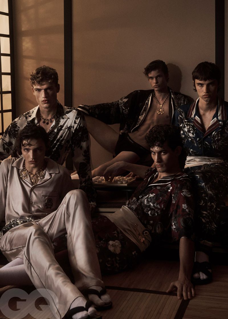 Five young adult men in patterned shirts and pants pose indoors, sitting on the floor in a fashion editorial shot with warm lighting.