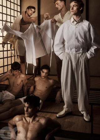 Fashion editorial featuring several shirtless adult male models posing indoors, wearing white bottoms or light-colored jackets in soft lighting.