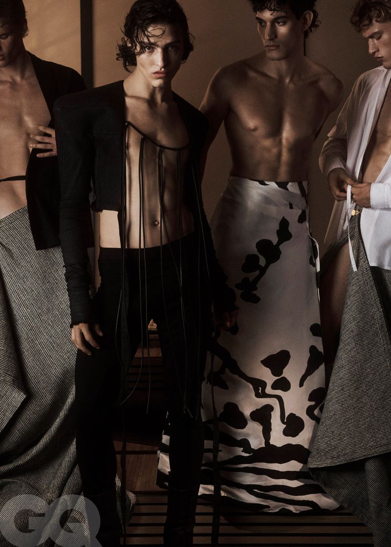 Fashion editorial featuring four adult male fashion models wearing tailored pieces and draped fabric in a neutral-toned studio setting.