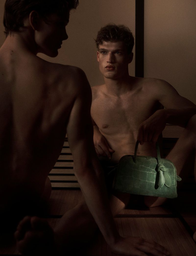 Two shirtless young men sit indoors in a dimly lit space with a green purse; one man faces forward, the other in profile.