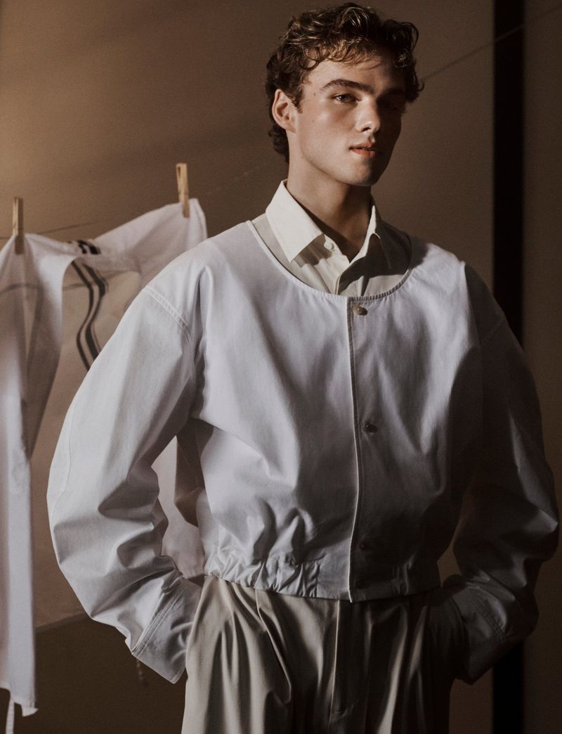 A young adult man poses in a white bomber jacket and light tan pants, standing indoors with clothing hung in the background.