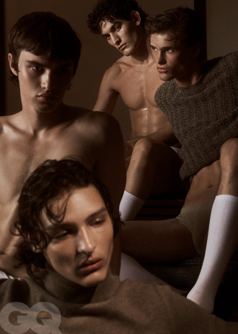 Four young adult males pose indoors in soft, muted lighting, fashion editorial; close-up portrait orientation shot with shallow depth of field.