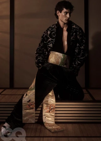 A male model kneels in a black embroidered jacket with an ornate gold brocade sash against warm studio panels.
