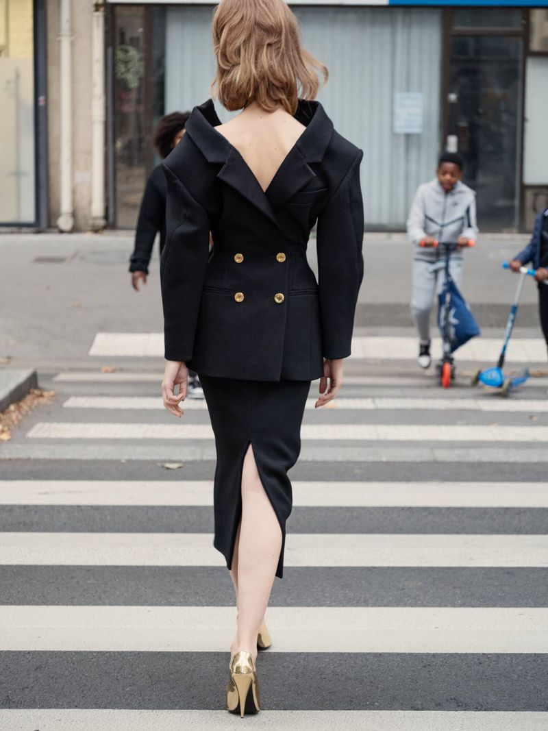 Woman in open-back blazer and pencil skirt walking on a crosswalk, other people and buildings in the background, outdoor setting.