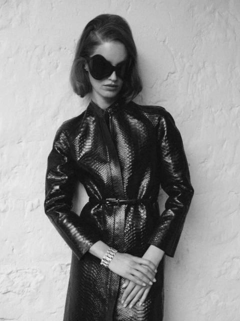 A woman with short dark hair wearing an all-black snakeskin-print outfit and large sunglasses stands against a textured off-white wall.