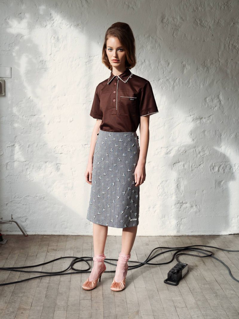 A stylish woman models a brown shirt and grey skirt in a fashion shot against a textured white wall with visible cables.